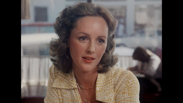 Close-up of a woman with styled wavy hair, wearing a yellow patterned blouse, sitting at a table in a cafe.