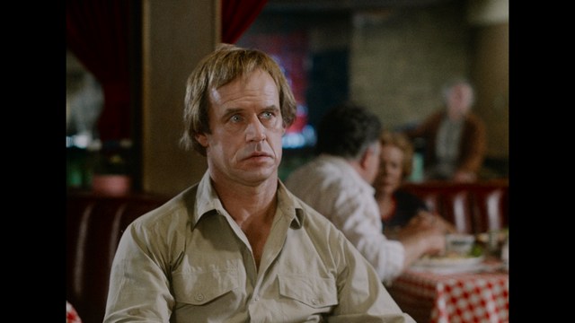 A man with short hair and a serious expression seated at a booth in a restaurant, with people and food in the background.