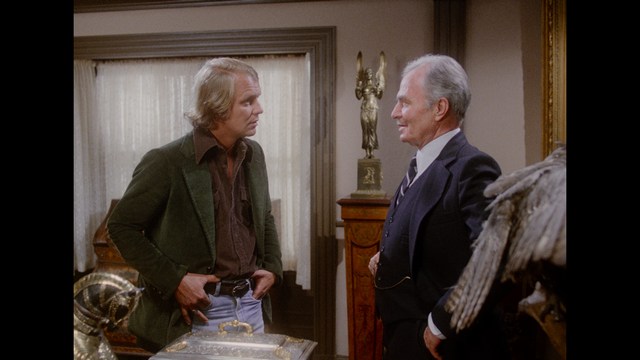 Two men engaged in conversation in an elegantly furnished room, featuring ornate decor and a statue in the background.