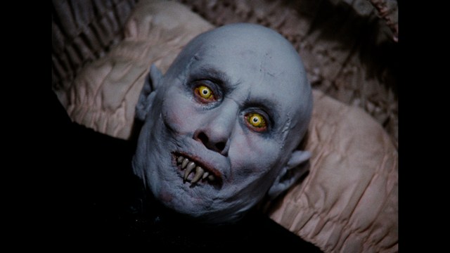 Close-up of a pale, bald figure with yellow eyes and sharp teeth, lying on a bed.