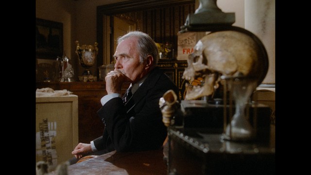 A character with large, white angel wings stands in a dimly lit room, wearing a black suit and looking at the camera with an intense expression.