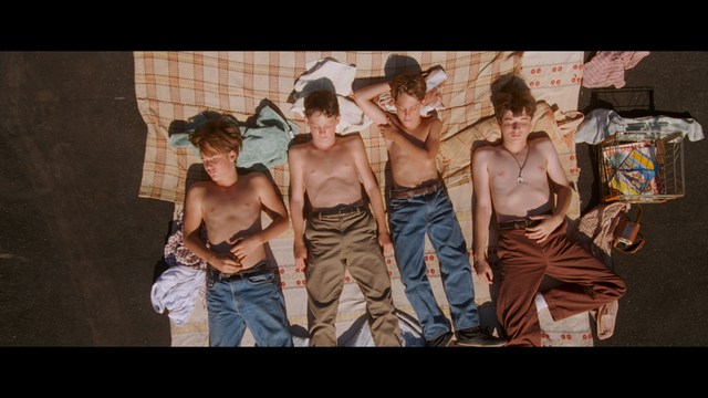 A still from a movie featuring a man standing on a boardwalk in front of an arcade with signs for Skeeball, saltwater taffy, and various attractions.