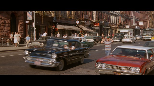 A man with a beard and mustache smiles warmly while standing on a boardwalk by the beach, with colorful flags overhead and a large building in the background.