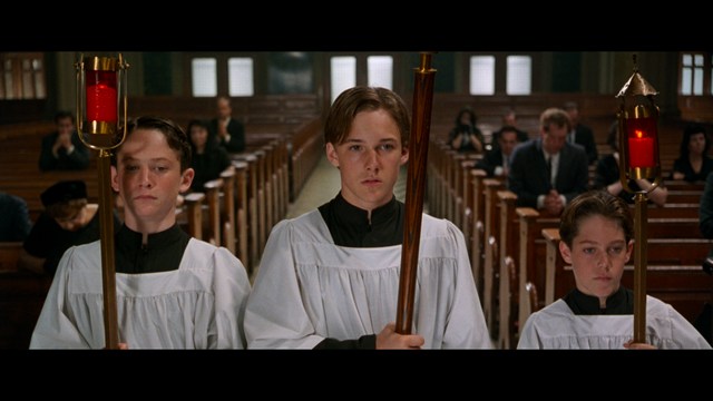 A serious-looking cardinal stands in front of a wooden backdrop with colorful posters related to Catholicism, holding a microphone at a press conference.