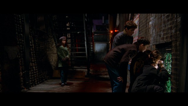 A scene from a movie featuring two young men sitting closely together in an airport waiting area, with one holding a box of popcorn.