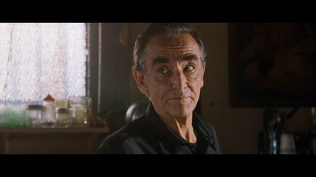 A woman with a pensive expression rests her chin on her hand, sitting at a table with kitchen appliances and items in the background.