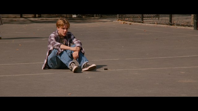A man in a white suit and hat stands at a doorway, smiling, with a suburban landscape in the background.
