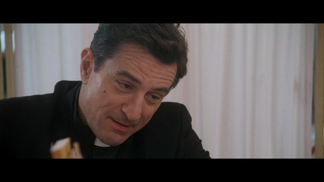 A man with large, feathered angel wings stands in a room, wearing a black suit and looking directly at the viewer.