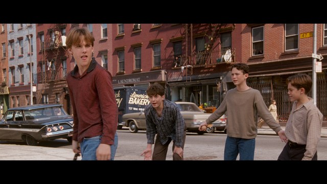 A close-up of a man with a concerned expression, with blurred figures in the background, set in a dimly lit interior.