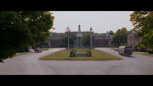 Close-up of a woman with long black hair sitting in a restaurant, looking concerned. A colorful painting is visible in the background, along with red bar stools.