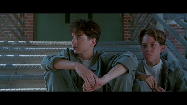 Two young men are looking at something off-camera, standing inside a building with bright lighting and seating areas visible in the background.