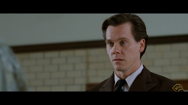 Close-up of a man with a serious expression, seated in a dimly lit area, with soft lighting from background lamps.