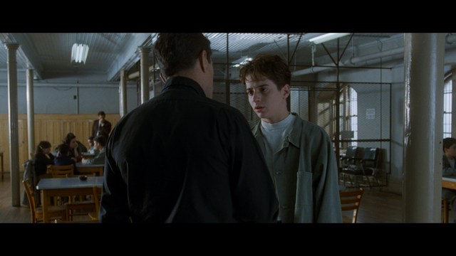 A close-up shot of a young man with short hair and a gray sweatshirt, looking intently at the camera, with a woman sleeping beside him in the background.