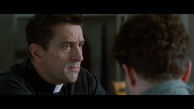 A woman seated at a restaurant table, thoughtfully resting her chin on her hand while reading a newspaper. Blurred figures of two people can be seen in the background.