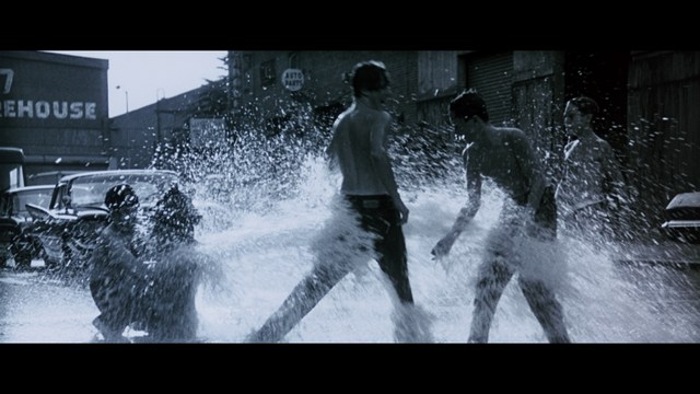 Two characters sitting at a table, one holding a small statue, with a surprised expression. The background features vibrant colors and patterns.