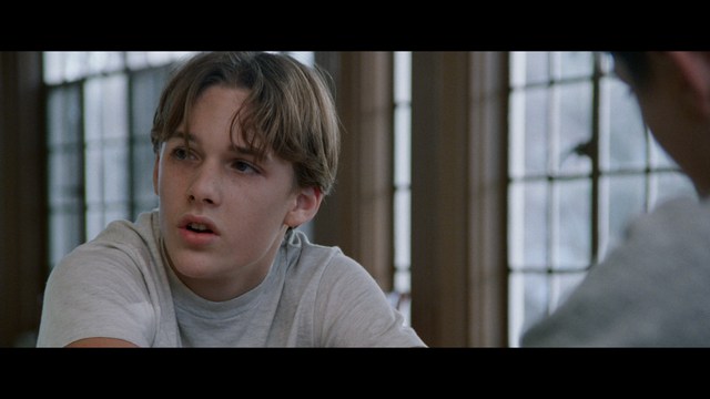 A serious-looking cardinal with a red hat and black robes sitting in a wooden chair in an office, surrounded by colorful posters and memorabilia.
