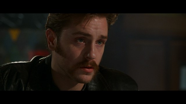 A shocked man with long hair and a baseball cap sitting in a bar, looking surprised.