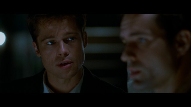 Close-up of an elderly woman in a wide-brimmed hat, with a serious expression. She is in an indoor setting with visible plants in the background.