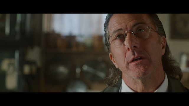 A scene from a film featuring two characters, one elderly woman holding a small object and speaking animatedly, and a young man listening intently, surrounded by lush greenery and plants in a greenhouse setting.