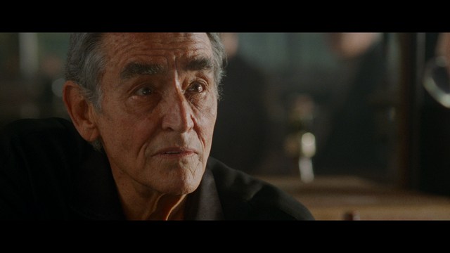 Close-up of a man with a concerned expression in a greenhouse setting, surrounded by various plants and soft lighting.