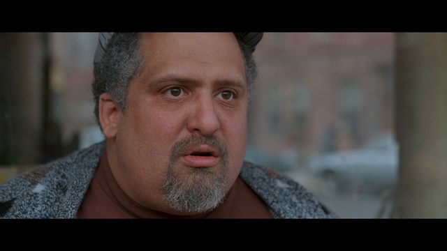 A close-up of an elderly man with a beard and white attire, appearing serious and contemplative, set against a colorful, futuristic background.