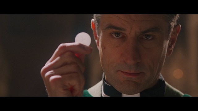 A close-up of an elderly man with a beard and white attire, appearing serious and contemplative, set against a colorful, futuristic background.