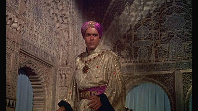 A man with large white wings stands in a room, wearing a dark suit, with a serious expression on his face. Bookshelves and curtains are visible in the background.