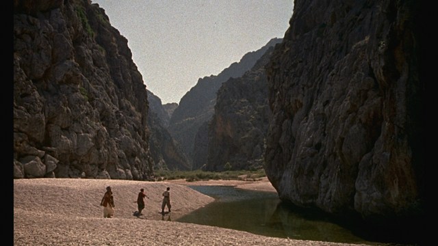 Scene featuring three characters on a roadside, discussing animatedly. A green landscape is visible in the background.