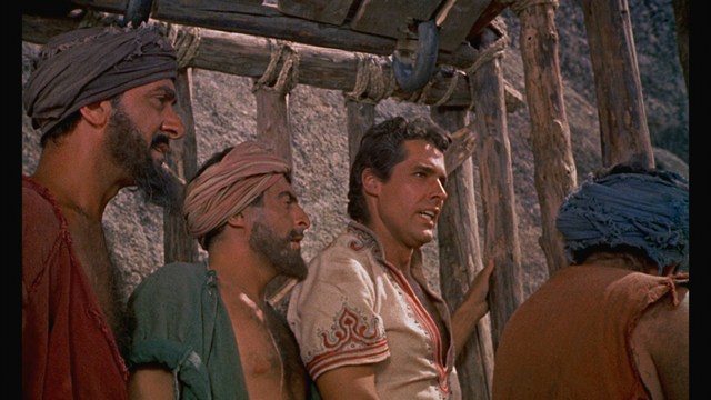 A scene from a film showing five characters in a bar setting, with a colorful background and a bartender visible in the back.