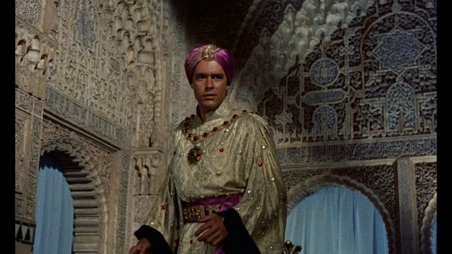 A man with large, feathered angel wings stands in a room, wearing a black suit and looking directly at the viewer.