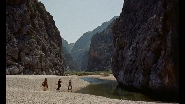 A scene from a movie featuring three characters standing on a bridge with a lush green landscape in the background.