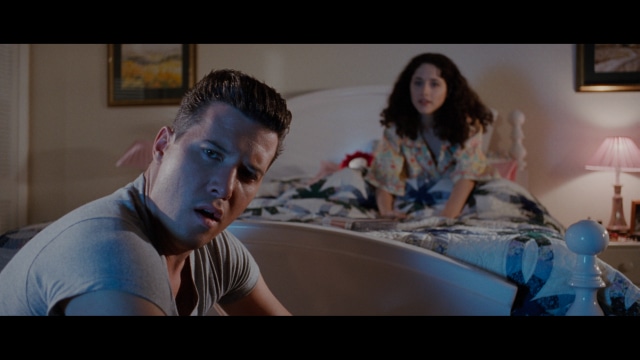 A close-up of a man with a concerned expression, with blurred figures in the background, set in a dimly lit interior.