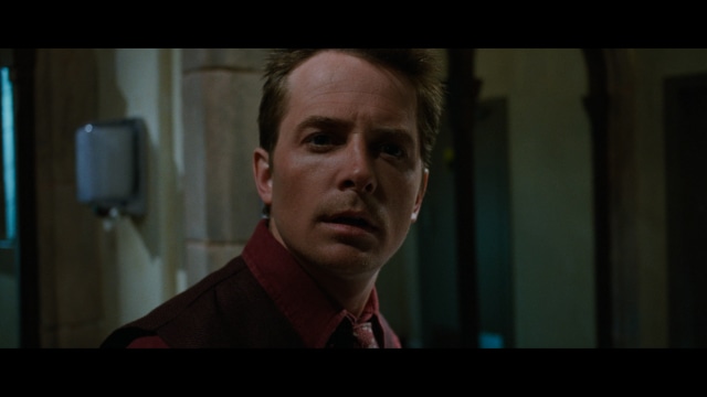 Two young men are looking at something off-camera, standing inside a building with bright lighting and seating areas visible in the background.