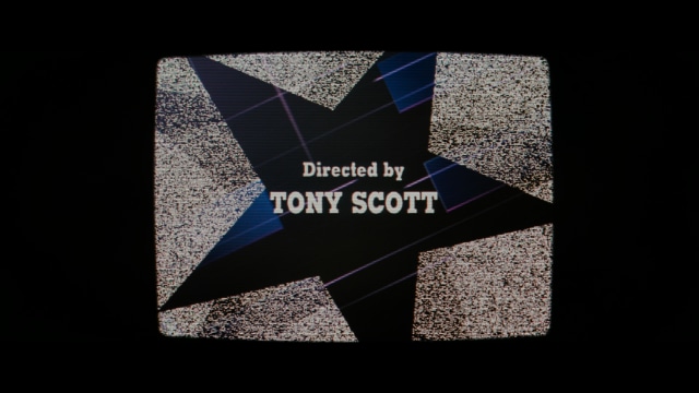 A still from a movie featuring a man standing on a boardwalk in front of an arcade with signs for Skeeball, saltwater taffy, and various attractions.