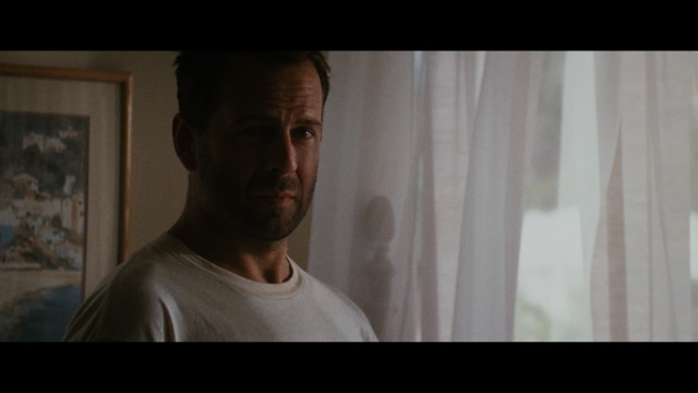 A close-up of a man with a concerned expression, with blurred figures in the background, set in a dimly lit interior.