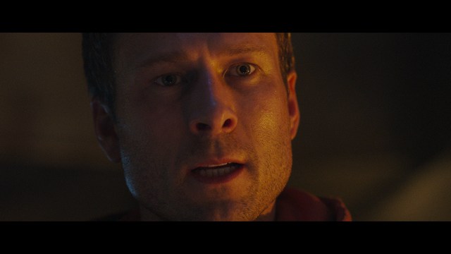A still from a movie featuring a man standing on a boardwalk in front of an arcade with signs for Skeeball, saltwater taffy, and various attractions.