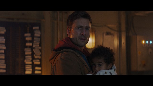 A man with a beard and mustache smiles warmly while standing on a boardwalk by the beach, with colorful flags overhead and a large building in the background.