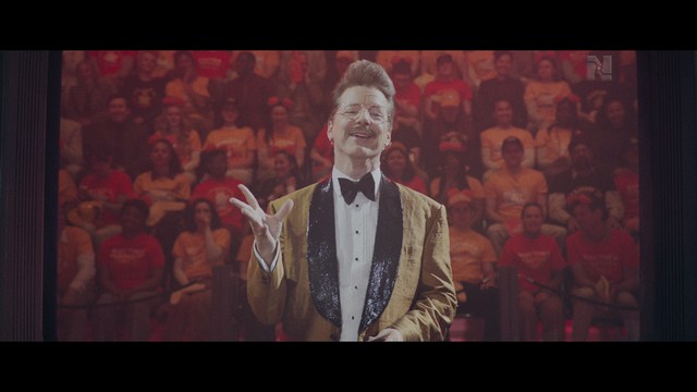 Statue of a smiling figure resembling Jesus, giving a thumbs up gesture, set against a colorful backdrop with signs related to Catholicism.