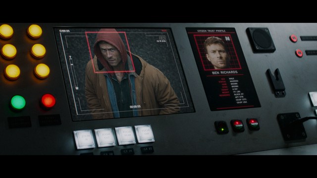 A woman with a pensive expression rests her chin on her hand, sitting at a table with kitchen appliances and items in the background.