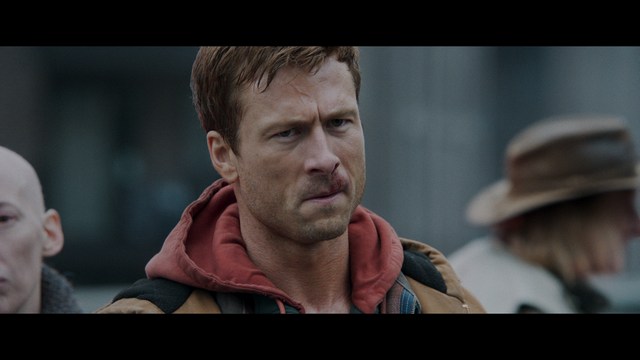 A man in a white suit and hat stands at a doorway, smiling, with a suburban landscape in the background.