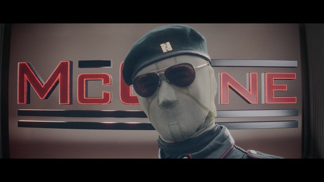 A man with large, feathered angel wings stands in a room, wearing a black suit and looking directly at the viewer.