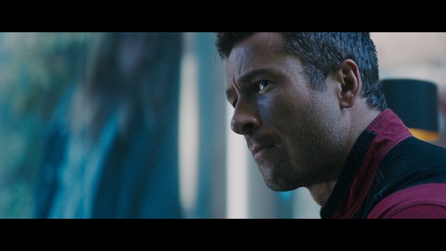 A close-up of a man with a concerned expression, with blurred figures in the background, set in a dimly lit interior.