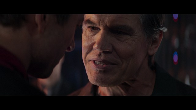 Two young men are looking at something off-camera, standing inside a building with bright lighting and seating areas visible in the background.