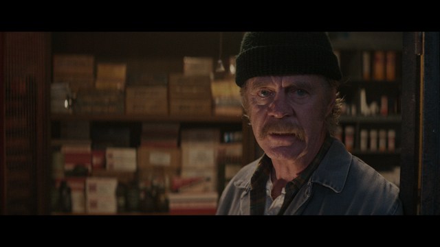 Close-up of a man with a serious expression, seated in a dimly lit area, with soft lighting from background lamps.