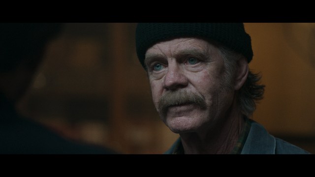 A close-up shot of a young man with short hair and a gray sweatshirt, looking intently at the camera, with a woman sleeping beside him in the background.