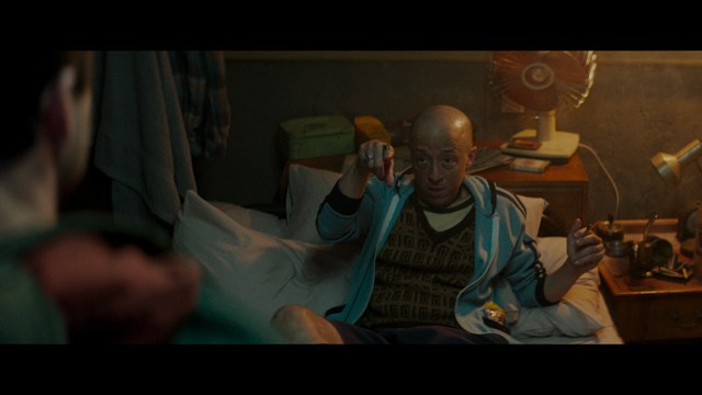 Two characters sitting at a table, one holding a small statue, with a surprised expression. The background features vibrant colors and patterns.