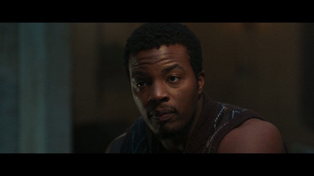 A scene featuring a man in a bishop's attire speaking at a podium surrounded by microphones, with a woman in a red dress beside him and a crowd of onlookers in the background.