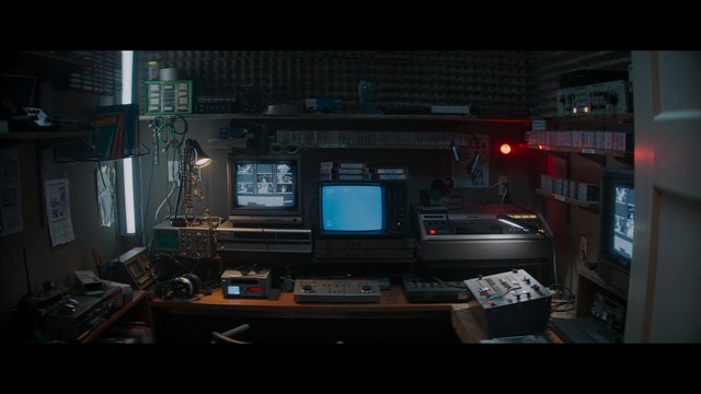 A tense scene featuring a woman with long hair and a serious expression, looking off to the side, while a man with short hair sits in the background, appearing relaxed and slightly turned away.