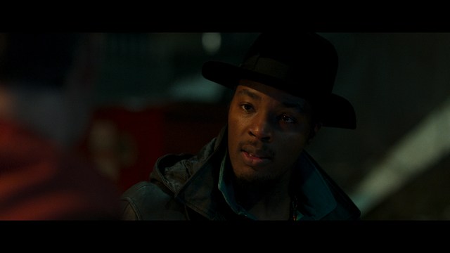Close-up of an elderly woman in a wide-brimmed hat, with a serious expression. She is in an indoor setting with visible plants in the background.