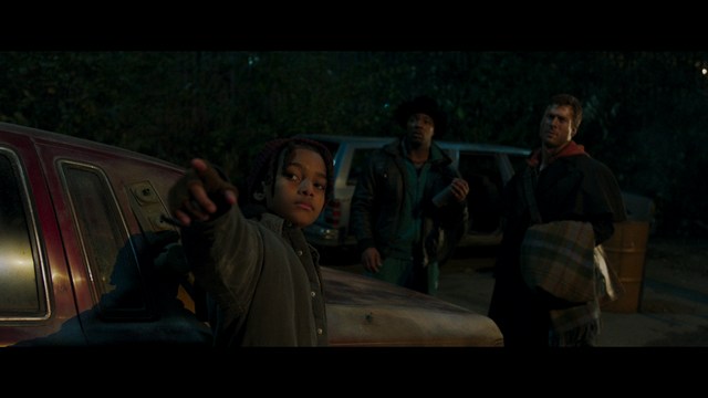 A scene from a film featuring two characters, one elderly woman holding a small object and speaking animatedly, and a young man listening intently, surrounded by lush greenery and plants in a greenhouse setting.